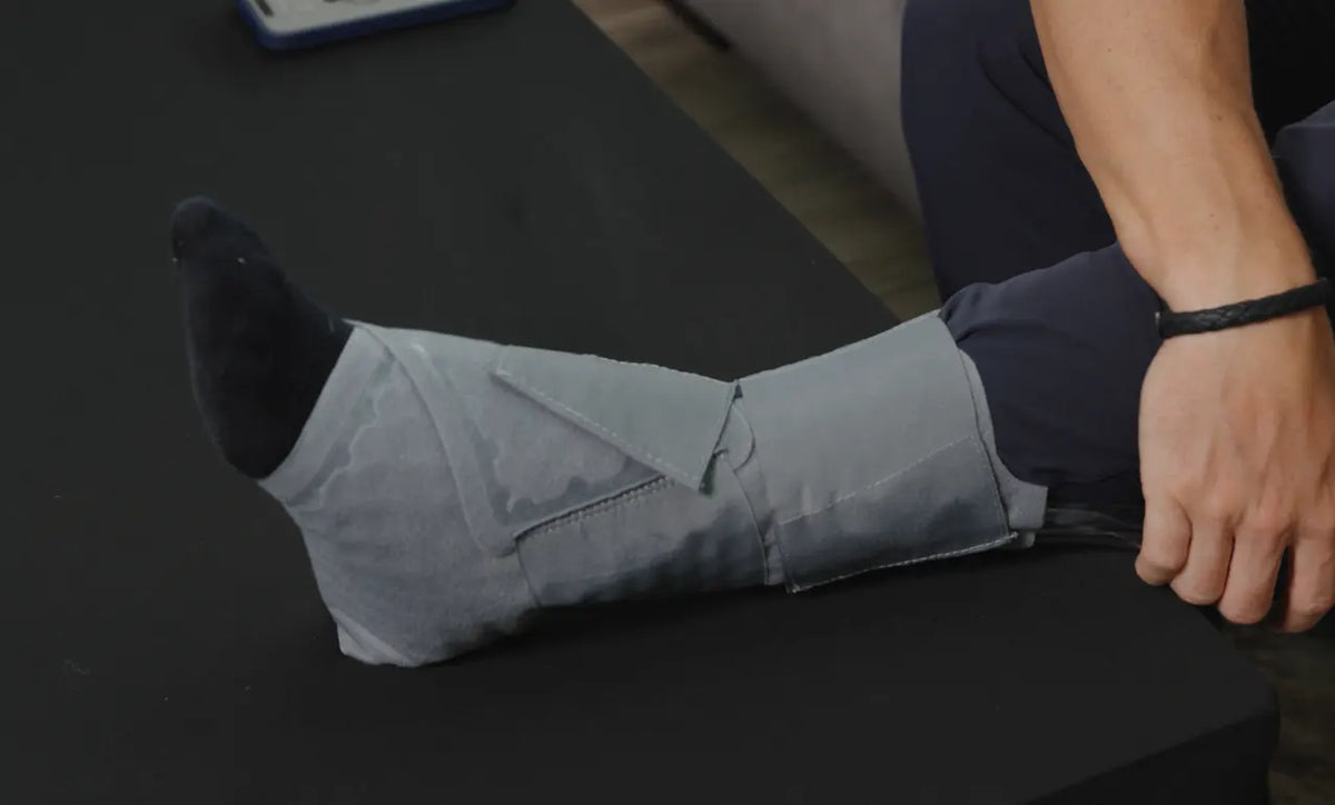 A person seated at a black table with one leg extended, wearing a light gray fabric leg wrap or cast support over a dark sock.